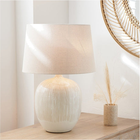 Greta Natural & Cream Textured Ceramic Table Lamp, 50cm, with ridged ceramic base and tapered fabric shade, shown on a wooden sideboard beside a woven mirror and dried grasses.