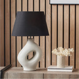 Ulla Monochrome Cream Organic Ceramic Table Lamp, 51cm, with sculptural cut-out base and black tapered shade, shown on a sideboard in a wood-panelled room.