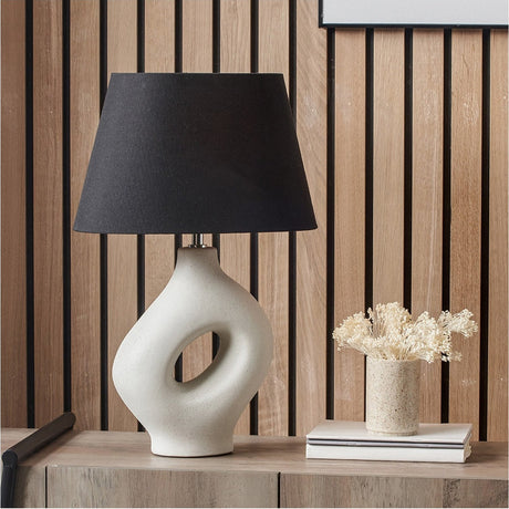 Ulla Monochrome Cream Organic Ceramic Table Lamp, 51cm, with sculptural cut-out base and black tapered shade, shown on a sideboard in a wood-panelled room.