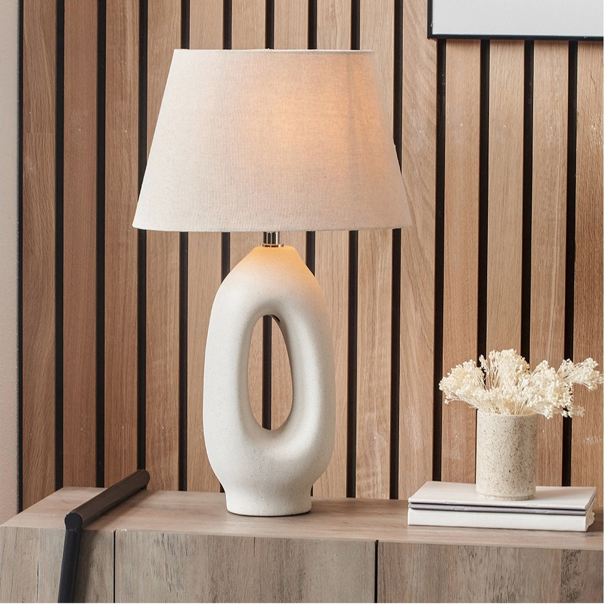 Laila Natural Beige Organic Tall Ceramic Table Lamp with a sculptural cut-out base and linen tapered shade, shown lit on a wooden sideboard against a slatted wood wall.