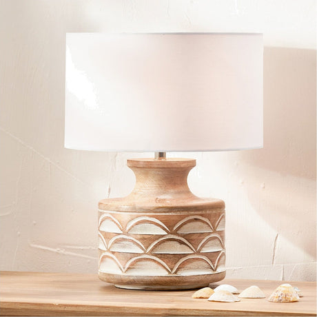 Kingsbury table lamp with white wash carved wood base featuring scalloped detailing and an ivory cotton drum shade, shown on a wooden surface with decorative shells.