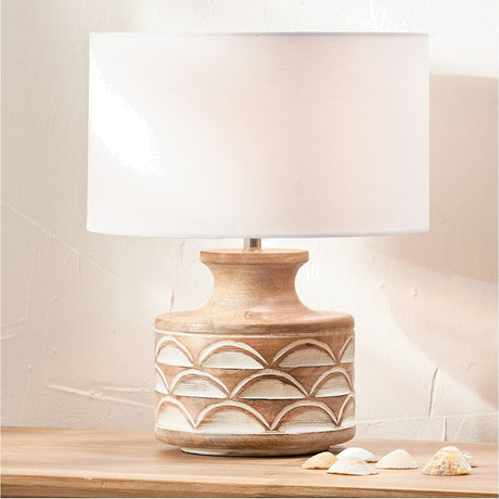 Kingsbury White Wash Carved Wood Table Lamp Base with white drum shade, arched carved pattern on rounded wood base, styled on a wooden surface with decorative shells.