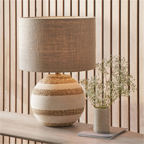 Talalla Table Lamp with round cream and natural sea grass striped base and grey linen drum shade, styled on a wooden console beside a vase of flowers.