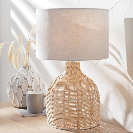 Caswell Rattan Cloche Table Lamp, 47cm, with a natural woven rattan cloche base and grey linen drum shade, shown on a wooden surface beside a vase and mug.