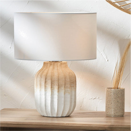 Amalia table lamp with a ribbed stoneware base in natural tan and white tones, paired with an ivory cotton cylinder shade, shown on a wooden sideboard.