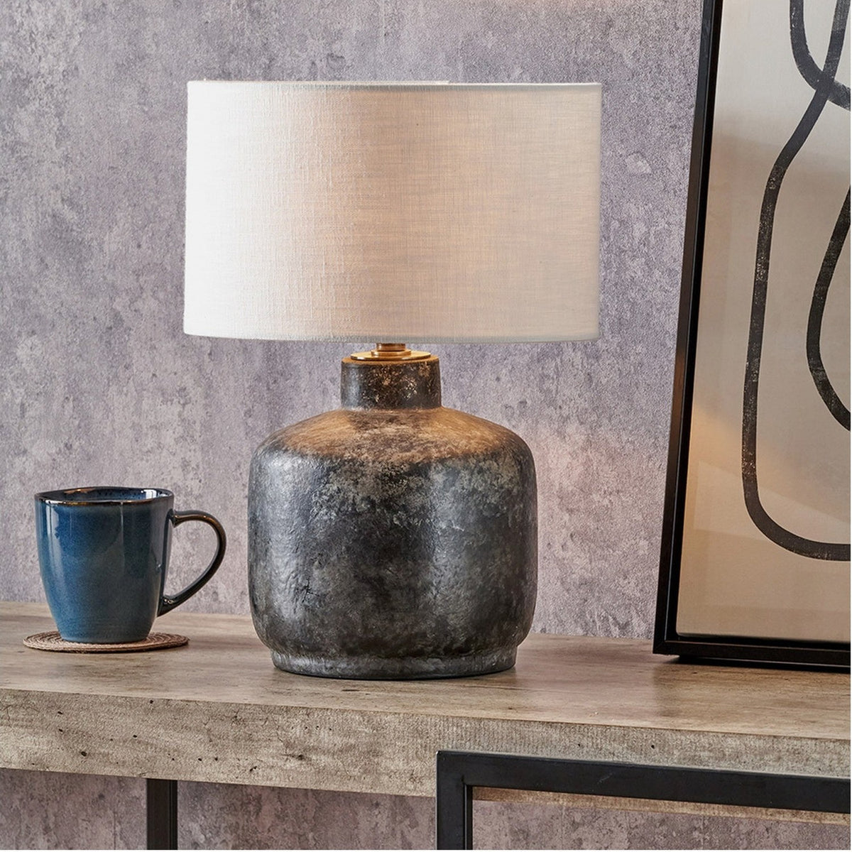 Vulcan table lamp with grey stoneware base in a textured volcanic-effect finish, paired with an oatmeal drum shade, styled on a console table beside a blue mug.