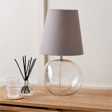 Hallie Table Lamp, 43cm, with a round clear glass base and tapered grey fabric shade, shown on a wooden sideboard beside a candle and reed diffuser.