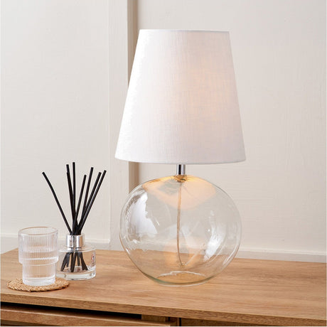 Hallie table lamp with clear round glass base and white tapered fabric shade, styled on a wooden sideboard beside a reed diffuser and glass tumbler.