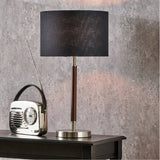 Gianni table lamp with black drum shade, brushed silver base and dark wood-effect stem, styled beside a vintage-look radio on a dark wood table.