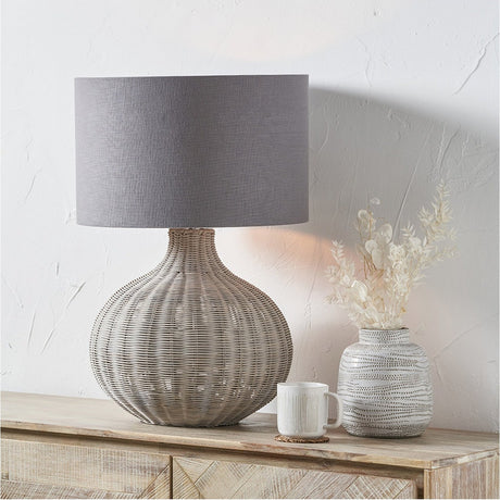 Allona Grey Wash Rattan Table Lamp with round woven base and grey drum shade, shown on a wooden sideboard beside a patterned vase and mug.