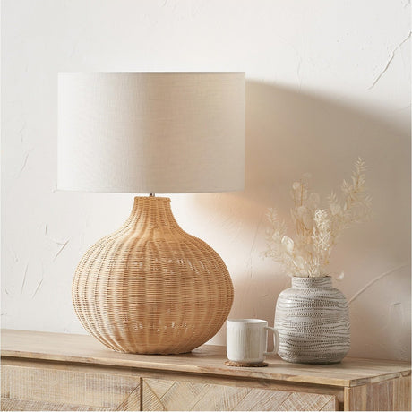 Allona Rattan Table Lamp - 70cm with a rounded woven natural rattan base and drum linen shade, styled on a wooden sideboard beside a patterned vase and mug.