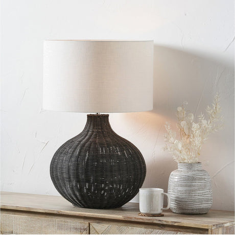 Allona Black Rattan Table Lamp, 70cm, with round woven base and white fabric drum shade, styled on a wooden sideboard beside a vase and mug.