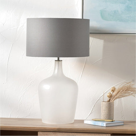 Tamsin Frosted White Glass Table Lamp, 59cm, with a bulbous frosted glass base and grey drum fabric shade, shown on a wooden sideboard beside a book and vase.