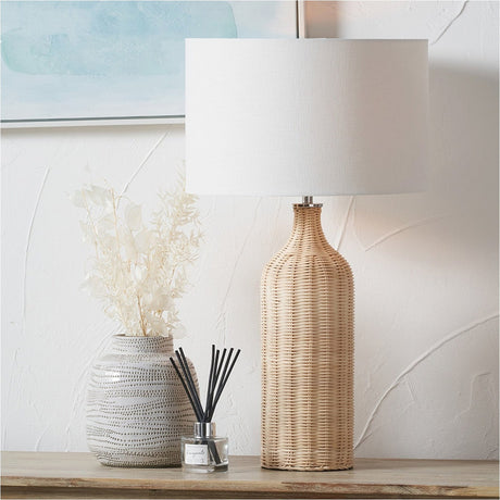 Anika Rattan Bottle Table Lamp, 62cm, with a natural woven rattan base and white drum shade, styled on a wooden console beside a patterned vase and dried foliage.