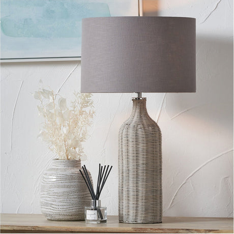 Anika Grey Wash Rattan Bottle Table Lamp, 62cm, with a woven rattan base and grey fabric drum shade, shown on a wooden surface beside a patterned vase and reed diffuser.
