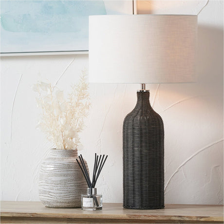 Anika Black Rattan Bottle Table Lamp, 62cm, with a woven black rattan bottle-shaped base and cylindrical white linen shade, shown on a wooden console beside a patterned vase and reed diffuser.