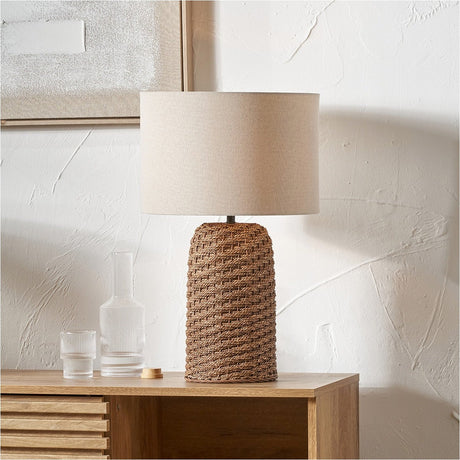 Andros Brown Woven Seagrass Table Lamp, 55cm, with a cylindrical woven base and beige linen drum shade, styled on a wooden sideboard against a textured white wall.