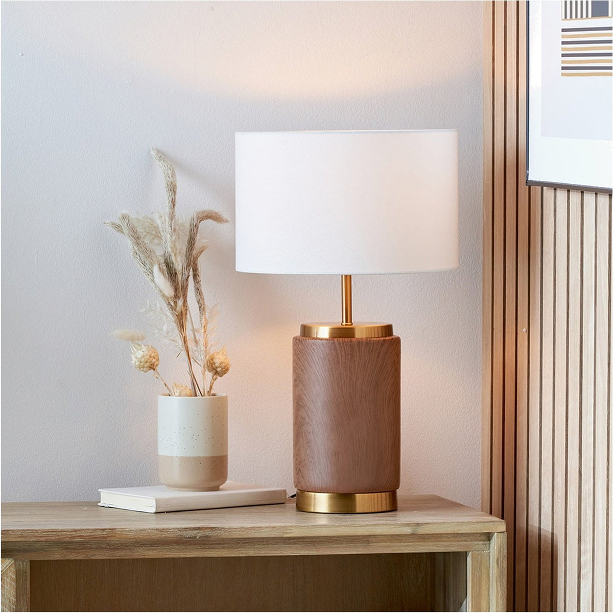 Carrara Wood Effect Ceramic Table Lamp, 48cm, with a wood-effect cylindrical ceramic base, brass fittings, and white fabric drum shade, styled on a wooden sideboard beside a vase of dried grasses.