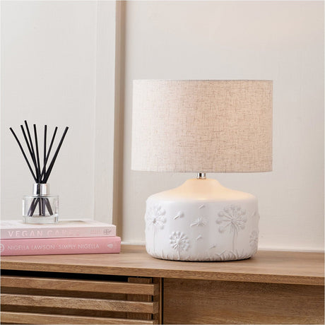 Flora White Small Floral Ceramic Table Lamp with a linen drum shade on a wooden sideboard, shown beside books and a reed diffuser.