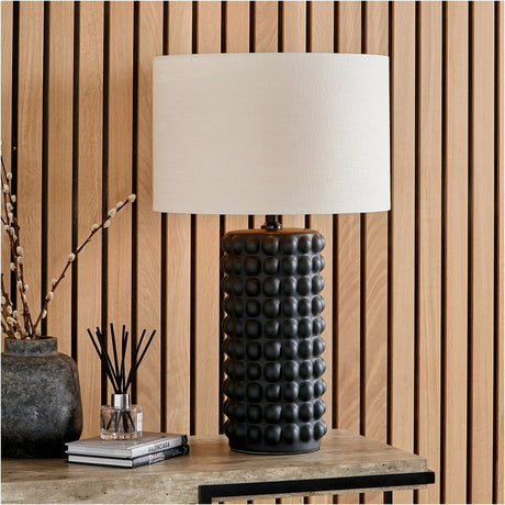 Sohan black bobbled ceramic table lamp with a cream drum shade, styled on a wooden console beside books and a vase against a slatted wood panel wall.