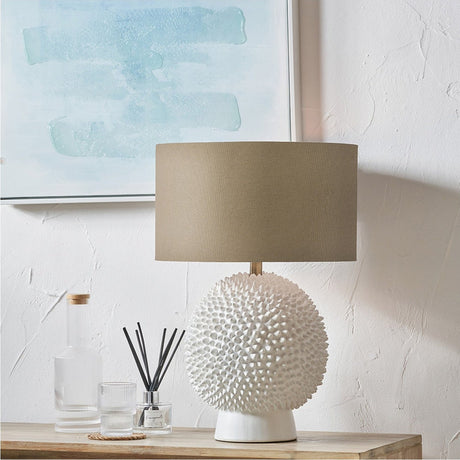 Wrenley Cream Ceramic Sea Urchin Table Lamp with Taupe Handloom Shade, textured spiky ceramic base and drum shade, shown on a wooden console beside glass bottles in a light-toned room.