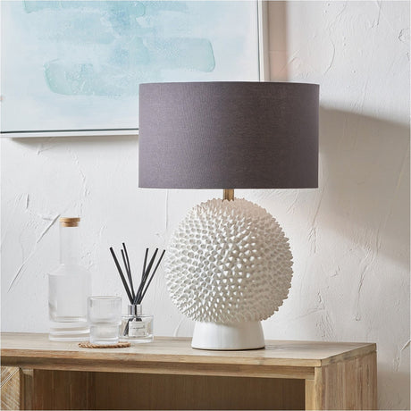 Wrenley Cream Ceramic Sea Urchin Table Lamp with textured spiked base and grey drum shade, styled on a wooden sideboard beside glass bottles.