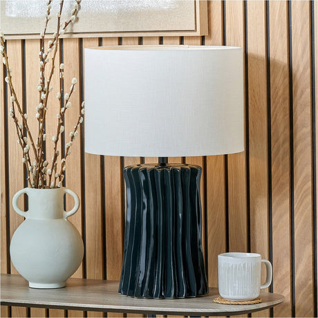 Devony Black Pleated Ceramic Table Lamp with white drum shade, styled on a wooden console table beside a textured vase and mug in a wood-panelled room.