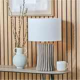 Devony Taupe Pleated Ceramic Table Lamp Base with white drum shade, styled on a console table beside a vase of branches and a mug against wood-panelled walls.