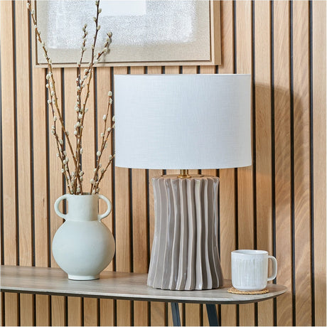 Devony Taupe Pleated Ceramic Table Lamp Base with white drum shade, styled on a console table beside a vase of branches and a mug against wood-panelled walls.