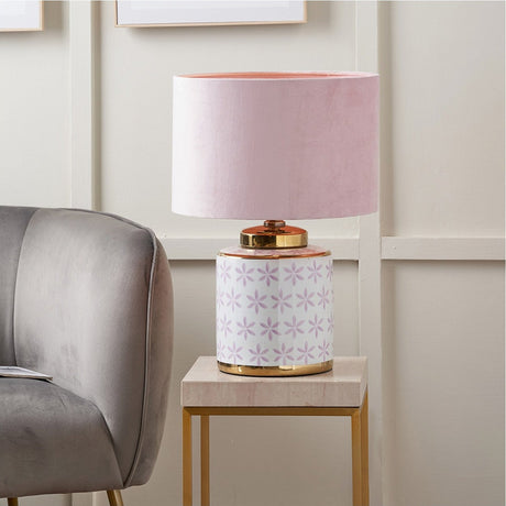 Thea Lilac & Gold Leaf ceramic table lamp with pink velvet drum shade, floral-patterned base and gold trims, shown on a marble side table in a living room setting.