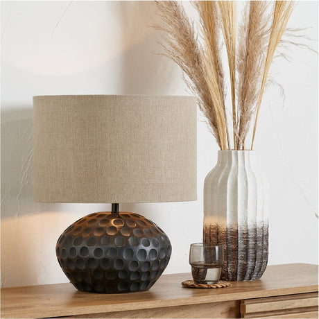 Dante Antique Black Mango Table Lamp, 42cm, with dimpled dark base and beige linen drum shade, styled on a wooden sideboard beside a ribbed vase of dried grasses.