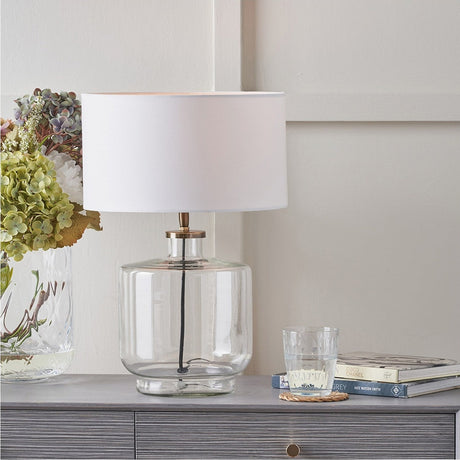 Josie Clear Glass & Antique Brass Table Lamp Base with white drum shade, styled on a grey sideboard beside hydrangeas, books, and a glass tumbler.