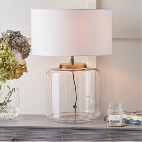 Josie Clear Glass & Antique Brass Large Table Lamp Base with white drum shade, shown on a sideboard beside hydrangeas in a living room setting.