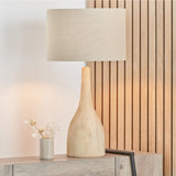 Corella Mango Curved Table Lamp Base, 54cm, in natural wood with a curved bottle-shaped body, shown with a beige drum shade on a wooden sideboard.
