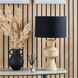 Koa Mango Contemporary Table Lamp Base, 43cm, with sculpted natural wood stacked geometric forms and black drum shade, styled on a table beside a black vase of dried foliage.