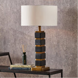 Maximo table lamp with matt black and antique brass cube-stacked base and white drum shade, styled on a dark side table beside books and a reed diffuser.