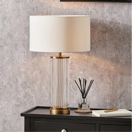 Ciara table lamp with fluted clear glass column and antique brass fittings, topped with a white drum shade, shown on a dark cabinet against a textured grey wall.