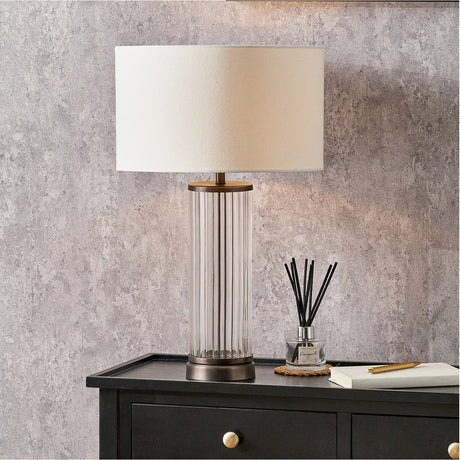 Ciara Clear Glass & Antique Bronze Table Lamp with ribbed cylindrical base and cream drum shade, shown on a dark sideboard in a living room setting.