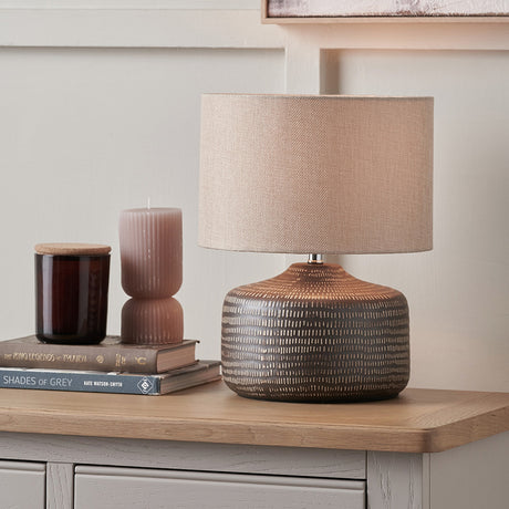 Kai Earth Brown Textured Ceramic Table Lamp, 34cm, with beige drum shade on a ridged brown ceramic base, styled on a wooden sideboard beside books and candles.