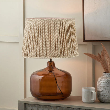 Corby Amber Glass Table Lamp, 58cm, with a rounded amber glass base and pleated taupe chevron fabric shade, shown on a sideboard in a living room setting.