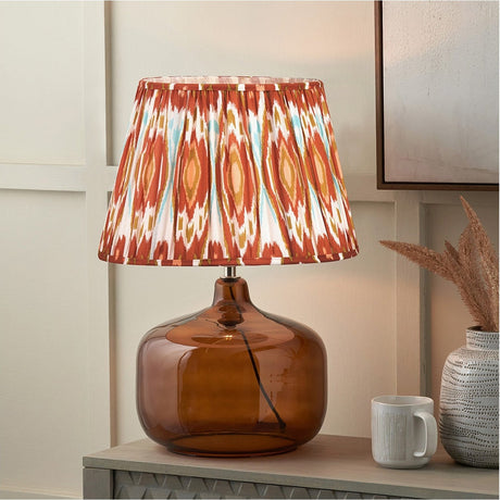 Corby Amber Glass Table Lamp, 61cm, with a bulbous amber glass base and pleated cinnamon Ikat print shade, shown on a sideboard beside a mug and vase.