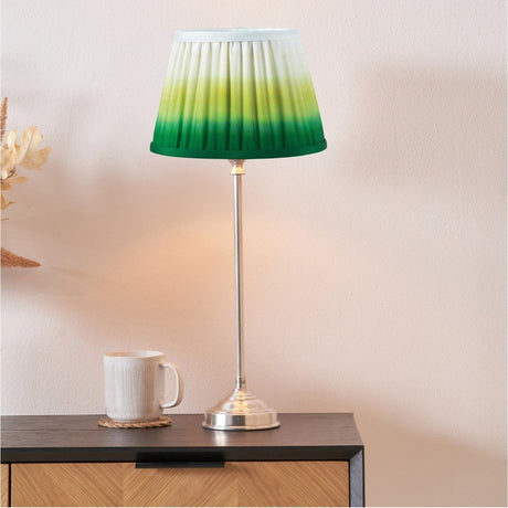 Auden silver metal stick table lamp, 52cm, with green ombre pleated fabric shade, shown lit on a dark wood sideboard beside a white mug in a living room setting.