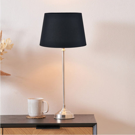 Auden table lamp with slim silver metal stick base and black tapered cotton shade, shown lit on a dark wooden sideboard beside a white mug.