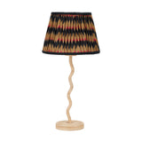 Palau Rattan Wave Table Lamp Base, 39cm, with a wavy natural rattan stem on a round ridged base, shown with a pleated black, tan and red patterned shade on a white background.