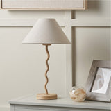 Palau Rattan Wave Table Lamp with natural wavy rattan base and neutral tapered fabric shade, styled on a sideboard beside a framed print and glass ornament.