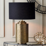 Zuri Large Antique Brass Pot Table Lamp with black drum shade, styled on a console table beside a decorative wire clock and metallic ornaments.