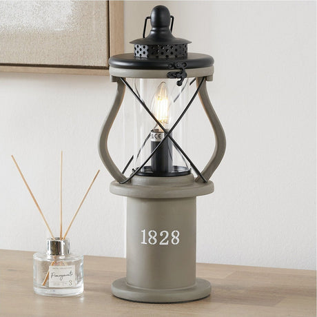Gibson Antique Grey Lantern Table Lamp, 41cm, with black lantern-style cage frame, glass shade and exposed bulb on a cylindrical base marked "1828", shown on a wooden table.