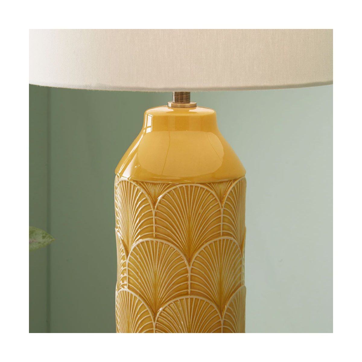 Close-up of the Bethan mustard yellow ceramic table lamp base with embossed art deco fan pattern, glossy neck, and cream fabric shade, set against a soft green wall.