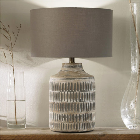 Galle Grey Wash Wood Textured Table Lamp with a carved ridged base and grey cotton drum shade, styled on a wooden shelf beside glass vases.