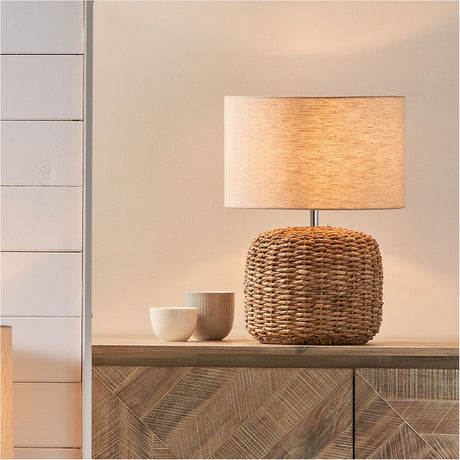 Acer Natural Woven Small Table Lamp with a round woven jute base and cream drum shade, shown lit on a wooden sideboard beside ceramic cups.